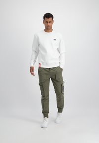 Sweatshirt blanc à col rond et logo, associé à un pantalon cargo vert olive avec poches et poignets élastiques, le tout complété par des baskets blanches.
