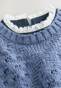 Suéter azul de punto con patrones texturizados, superpuesto a una blusa azul oscura con borde de encaje blanco en el escote.