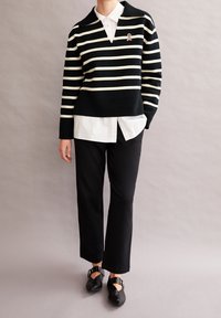 Personne portant un pull rayé noir et blanc par-dessus une chemise blanche, un pantalon noir et des chaussures à boucle noires, se tenant contre un fond gris.