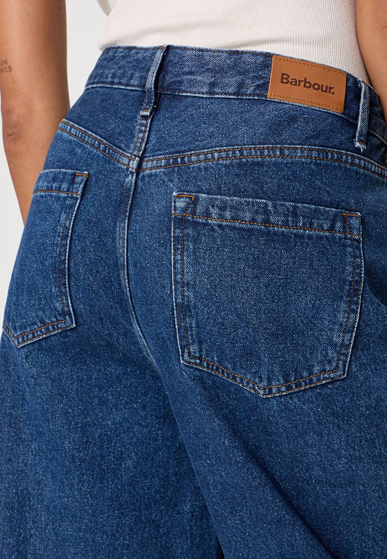 Person, der blaue Jeans mit Gesäßtaschen und einem braunen Lederetikett der Marke Barbour am Bund trägt.