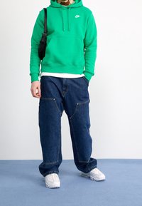 Grön hoodie med känguruficka och vitt logo. Mörkblå baggy denimjeans med stora framfickor. Vita sneakers. Enfärgad bakgrund.