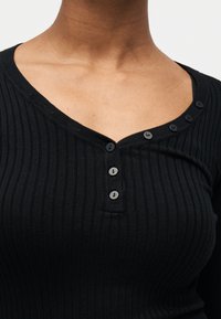 Gros plan sur une personne portant un haut noir côtelé avec un col en V et cinq petits boutons noirs le long de l'encolure et de la poitrine.