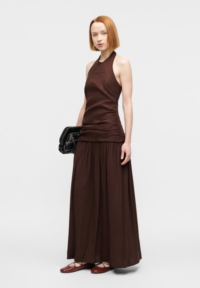 Καφέ τοπ με λουρί στο λαιμό, με εφαρμοστό μπούστο και συγκεντρωμένη μέση, που συνδυάζεται με φούστα σε μήκος μέχρι τους αστραγάλους, με πιέτες και ρέον ύφασμα· φορεμένο με κόκκινα μπαλαρίνια.