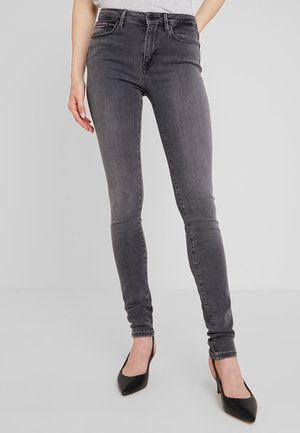 Kvinde iført tætsiddende, højttaljede grå jeans kombineret med sorte spidse hæle, stående foran en ensartet lys baggrund.