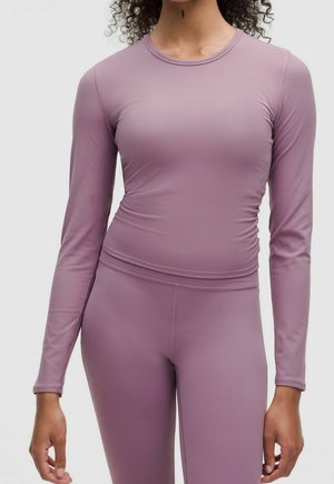 Femme portant un haut moulant à manches longues mauve et des leggings taille haute assortis, debout devant un fond clair uni.