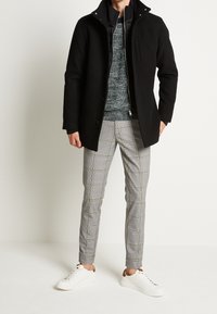 Manteau noir avec un col montant, pull en laine gris texturé et pantalon à carreaux gris. Des baskets blanches avec des accents marron complètent l'ensemble.