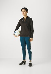 Zwart sportief shirt met lange mouwen en een halflange rits, gecombineerd met teal geklede broek met witte zijstrepen, terwijl hij een meerkleurige voetbal vasthoudt.