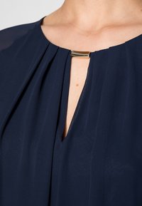 Mörkblå blus med en plisserad design, V-ringning och en metallisk detalj vid kragen, som visar en mjuk och flödande tygtextur.