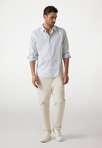 Camicia a righe blu chiaro con colletto button-down, abbinata a pantaloni color crema e sneakers bianche. La camicia ha le maniche arrotolate.