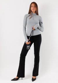 Top zip grigio chiaro a coste con maniche lunghe, dotato di fori per il pollice, abbinato a pantaloni neri a palazzo e tacchi neri a punta, mentre tiene un'etichetta.