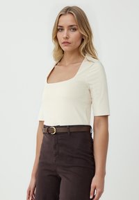 Crème korte mouwen top met een vierkante halslijn, gecombineerd met hoge donkerbruine broeken en een bijpassende riem met een gouden ronde gesp.
