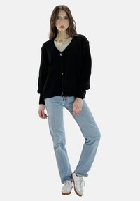 Cárdigan negro de punto con cuello en V, mangas largas y cierre de botones en la parte frontal, combinado con vaqueros de un azul claro y zapatillas blancas.
