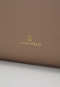 Surface en cuir marron texturée avec logo embossé doré et texte ANNA FIELD sur un sac à main ou un portefeuille.