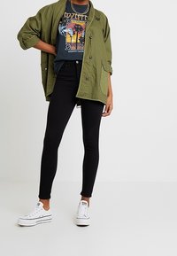 Olive green overshirt with rolled sleeves, graphic black t-shirt, black skinny jeans, and white sneakers with black accents.