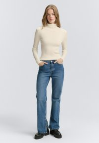 Pulover crem cu guler înalt, din tricot cu model ribbed, blugi evazați albaștri cu talie înaltă și cizme negre cu talpă platformă. Design simplu, fără modele sau ornamente vizibile.