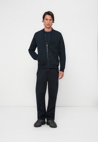 Sweatshirt zippé bleu marine avec un col rond, associé à un pantalon cargo assorti avec plusieurs poches et des chaussures noires texturées.