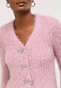 Gaiši rozā pūkaina kardigāna ar trim dekoratīvām pērlītēm rotātām lentēm. Audumam ir teksturēta, pūkaina izskata virsma ar maigu spīdumu.