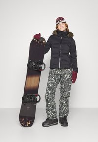 Čierna vypchatá bunda, vzorované snehové nohavice v zelenej a krémovej farbe, snowboardové okuliare a snowboard s čiernymi viazaniami a dreveným vzorom.