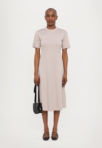 Vaaleanbeige polvipituinen mekko, jossa on lyhyet hihat, pyöreä pääntie ja näkyvät saumat; käytetty mustan olkalaukun ja ruskeiden kenkien kanssa.