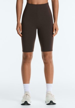 Kvinde iført mørkebrune tætsiddende biker shorts, hvide ankelsokker og hvide sneakers, stående mod en ensfarvet lys baggrund.