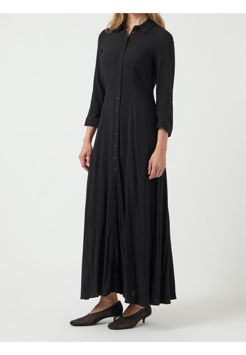 Femme portant une robe noire à manches longues, boutonnée, longueur cheville, associée à des chaussures noires à talons bas, debout sur un fond uni.