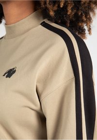 Sudadera beige con cuello alto, con rayas laterales en negro y crema y un gráfico de un oso negro en el pecho. Textura de tela suave.