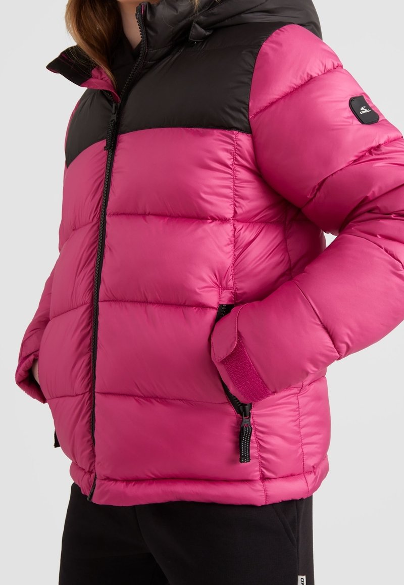 Veste d'hiver matelassée rose et noire avec fermeture éclair frontale, poches latérales et un écusson logo sur la manche gauche, portée avec les mains dans les poches.