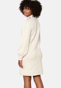 Abito a maglia beige con maniche a sbuffo lunghe e un collo alto, caratterizzato da dettagli con bottoni. Tessuto morbido, vestibilità affusolata e lunghezza sopra il ginocchio.