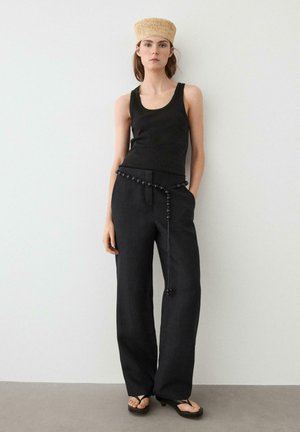 Kvinde iført sort tanktop, sorte bukser med vide ben og perlebeslået bælte, stråhat og sorte thong-sandaler står op ad en hvid væg.