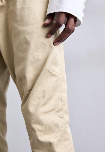 Pantaloni di cotone beige con una texture liscia e un dettaglio con tasca laterale, con una mano appoggiata sulla coscia.