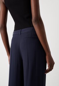 Personne portant un pantalon large à rayures bleu marine et un haut noir sans manches, vue de dos sur un fond uni.