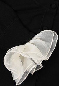 Crna pletenina s bijelim prozirnim volanima na rukavu, s glatkom teksturom i delikatnim valovitim rubovima.