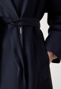 Gros plan d'une personne portant un manteau bleu marine foncé avec une ceinture nouée et la main reposant dans une poche latérale.