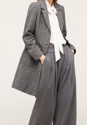 Femme portant un blazer gris à carreaux, une blouse blanche avec un nœud papillon et un pantalon gris taille haute à jambes larges, les mains dans les poches.