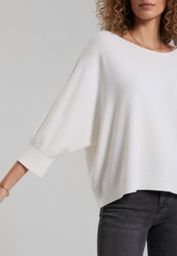 Weißer übergroßer Pullover mit Fledermausärmeln, gerippten Bündchen und abgerundetem Saum. Weiche Textur, minimalistisches Design, kombiniert mit dunklen Jeans.