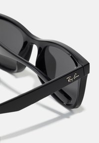 Ochelari de soare Ray-Ban negri cu un cadru lucios, formă rectangulară, lentile închise la culoare și un accent logo auriu pe bradă.