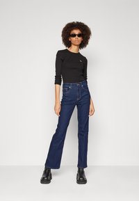 Svart langermet topp med subtilt logo, kombinert med mørkeblå rette jeans. Modellen har på seg klumpete svarte støvler og smale solbriller.
