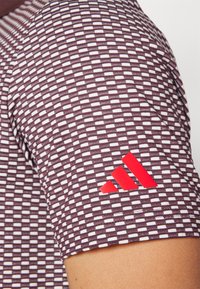Krátký rukáv sportovního trička v tmavě vínové barvě s mřížkovaným vzorem. Obsahuje červené logo Adidas na rukávu. Hladká, prodyšná tkanina.