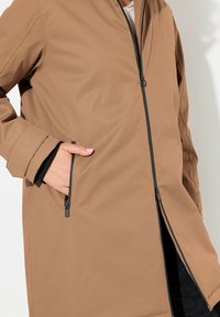 Veste marron mi-longue avec une fermeture éclair à l'avant, des poches latérales et des accents noirs sur la fermeture éclair et les poignets. Tissu texturé avec une finition lisse.