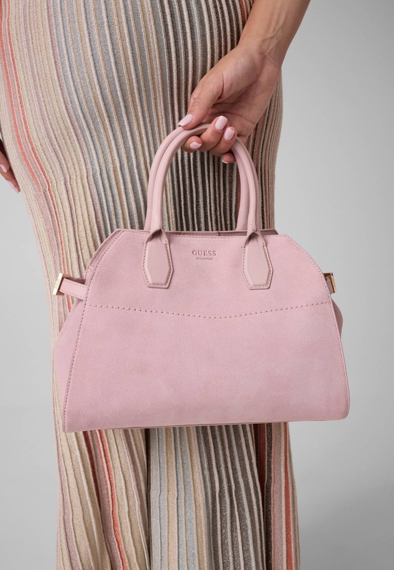 Main tenant un sac à main rose Guess avec double poignée sur un fond de jupe plissée multicolore à rayures verticales.