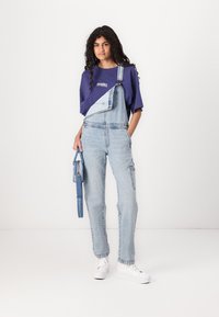 Salopette en denim bleu clair avec un devant croisé, portée avec un t-shirt à manches courtes violet. Accessoire : sac bandoulière bleu. Baskets blanches.