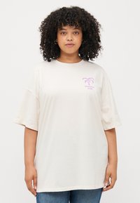 Kremfarget oversized t-skjorte med korte ermer, med en lilla trykk av palmer og teksten "SUNSHINE CLUB" under. Myk stofftekstur.