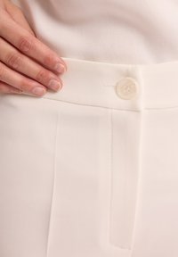 Białe dopasowane spodnie z płaskim przodem, z pojedynczym zapięciem na guzik i pionowymi szwami, wykonane z gładkiego materiału.