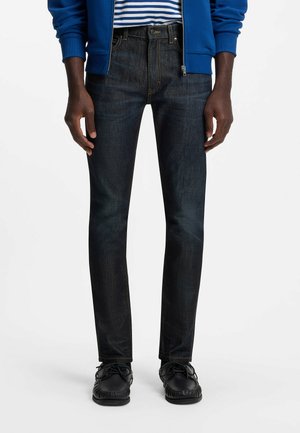 Uomo che indossa jeans slim fit blu scuro, scarpe nere con lacci, felpa blu con zip e camicia a righe blu e bianca, in piedi con le mani rilassate.