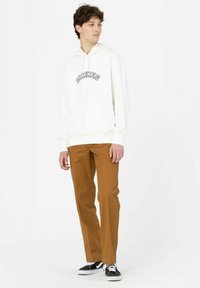 Weißer Kapuzenpullover mit "DICKIES"-Logo, kombiniert mit braunen Hosen und schwarzen Sneakers. Glattes Material, lässige Passform und Kängurutasche.