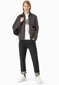 Giacca a vento grigia con tasca a toppa, maglietta bianca con grafica a girocollo, pantaloni in denim scuro e sneakers bianche con dettagli neri.