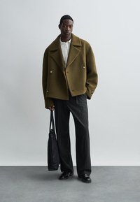 Khaki dvojradový vlnený kabát s veľkými klopami, spárovaný s pruhovanými nohavicami, čiernymi topánkami a čiernou kabelkou.