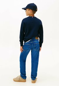 Marineblå langærmet skjorte, blå denim jeans og beige sneakers. Skridtet har små røde og hvide detaljer ved halsen.