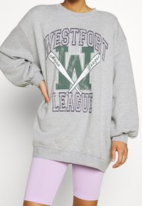 Šedá bavlněná oversized mikina s grafikou "WESTFORT LEAGUE", která má zelené a fialové detaily a raglánové rukávy, v kombinaci s levandulovými šortkami.