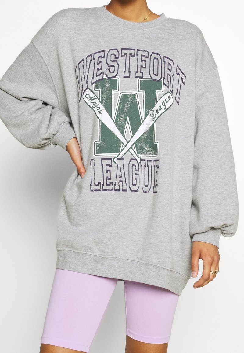 Šedá bavlněná oversized mikina s grafikou "WESTFORT LEAGUE", která má zelené a fialové detaily a raglánové rukávy, v kombinaci s levandulovými šortkami.